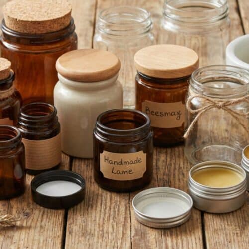 Mehrere leere und gefüllte Tiegel aus Braunglas, Klarglas, Keramik und Metall stehen auf einem Holztisch, umgeben von Bienenwachs, getrockneten Kräutern und einem Mörser – Materialien zur Salbenherstellung.