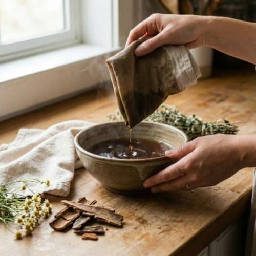 Eine Nahaufnahme auf einem rustikalen Holztisch: Eine Hand hebt ein nasses, naturfarbenes Leinentuch aus einer Keramikschüssel, die mit einem sehr dunklen, konzentrierten Kräuterauszug gefüllt ist. Dunkle Tropfen fallen vom Tuch zurück in die Schüssel. Daneben liegen getrocknete Rindenstücke. Das Bild zeigt das Tränken des Tuches für einen Umschlag.