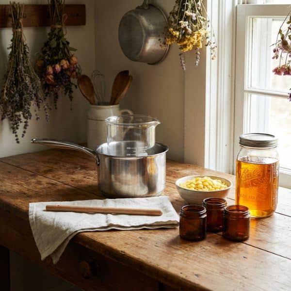 Ein stimmungsvolles Stillleben auf einem rustikalen Holztisch, das die Vorbereitung für die Salbenherstellung zeigt. Ein großes Glas mit gelben Bienenwachs-Pastillen und einem Holzlöffel steht neben einer Flasche mit goldenem Ölauszug. Mehrere leere, offene Braunglas-Tiegel mit Deckeln stehen sauber aufgereiht bereit zum Abfüllen. Weiches Tageslicht fällt durch ein Fenster auf die Szenerie.